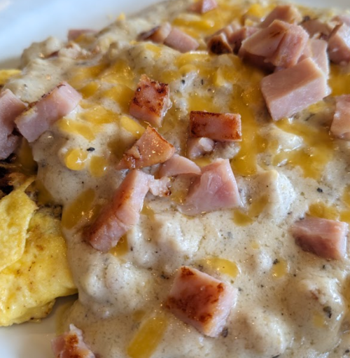 A close-up of a plate featuring an omelet topped with creamy gravy, diced ham, and melted cheese.