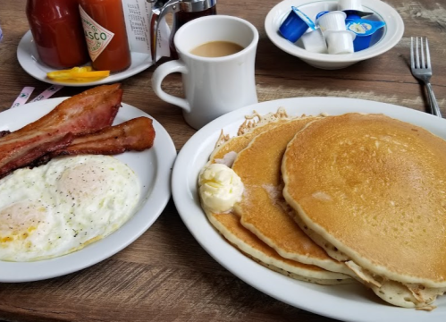 A plate of pancakes with butter, alongside eggs and bacon, with a cup of coffee and creamers in the background.