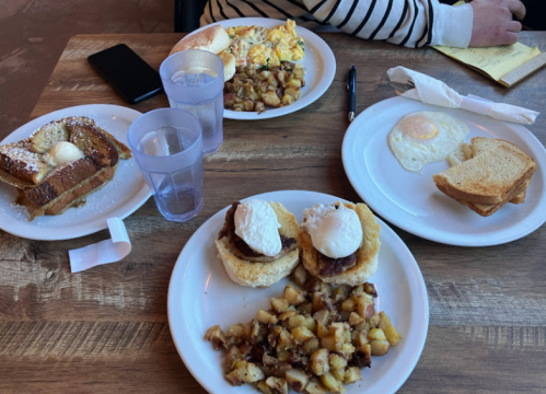 A table with breakfast dishes: French toast, eggs, potatoes, and biscuits with poached eggs, alongside drinks and utensils.