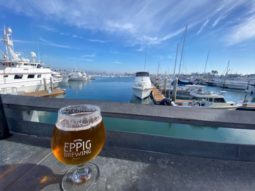 A glass of beer sits on a table overlooking a marina filled with boats under a clear blue sky.