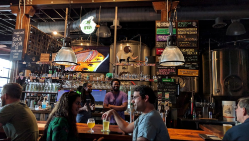 A lively bar scene with patrons enjoying drinks, a bartender serving, and brewing equipment in the background.