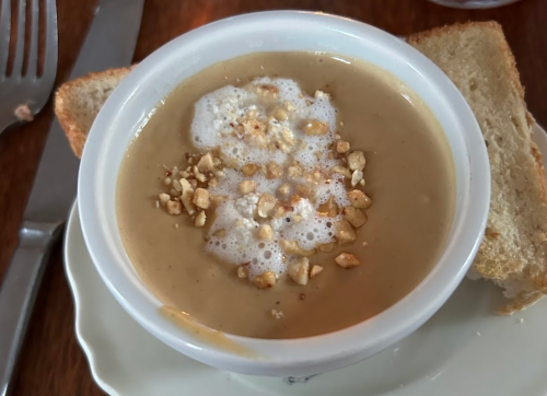 A bowl of creamy soup topped with foam and crushed nuts, served with two slices of bread on the side.