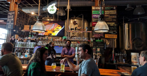 A lively bar scene with patrons enjoying drinks, a colorful menu, and brewing equipment in the background.