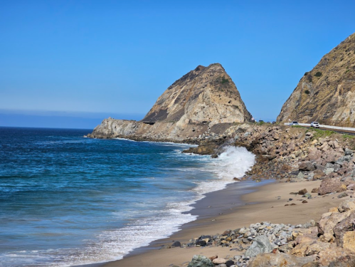 A rocky coastline with waves crashing on the shore, featuring a large cliff under a clear blue sky.