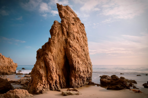 A tall, jagged rock formation stands on a sandy beach, with gentle waves lapping at its base under a blue sky.
