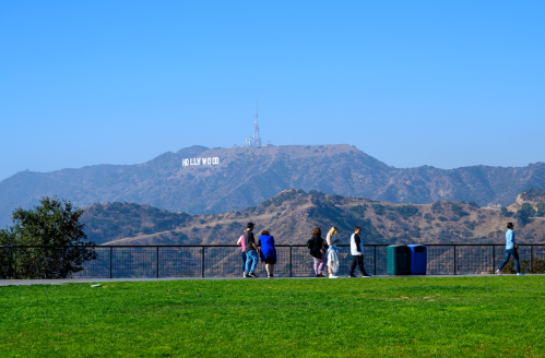 A group of people walks along a path with the Hollywood sign visible on a distant hill under a clear blue sky.