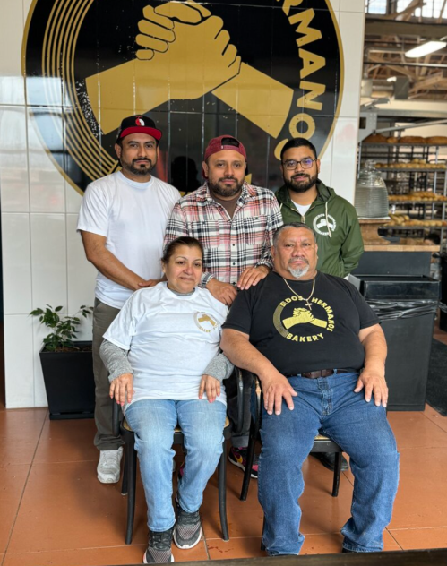 A group of five people, two seated and three standing, in a bakery setting with a logo in the background.