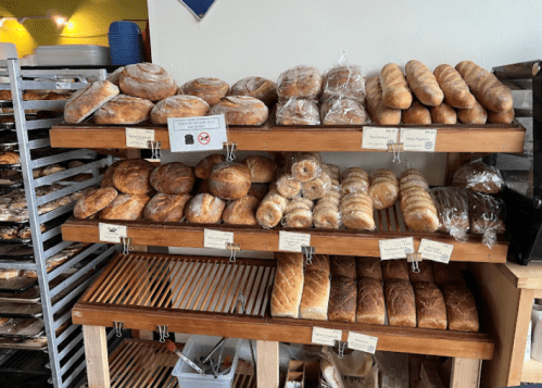 A display of various freshly baked breads on wooden shelves in a bakery.