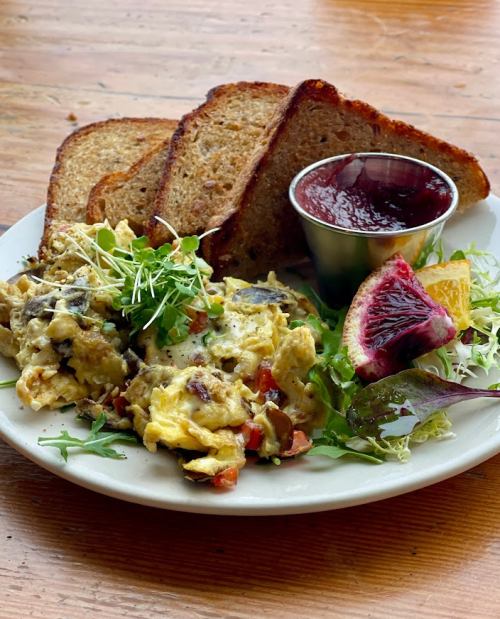 A plate with scrambled eggs, mixed greens, toast, and a small cup of jam.