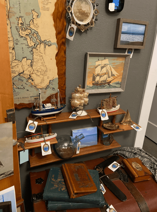 A display of model ships, nautical art, and vintage books on a wooden shelf against a textured wall.