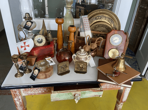 A table displaying various vintage items, including clocks, lamps, and decorative pieces, in a bright room.