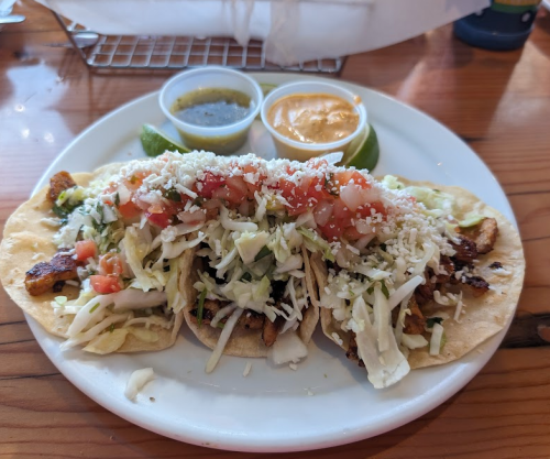 A plate of three tacos topped with lettuce, tomatoes, cheese, and served with two sauces and lime wedges.