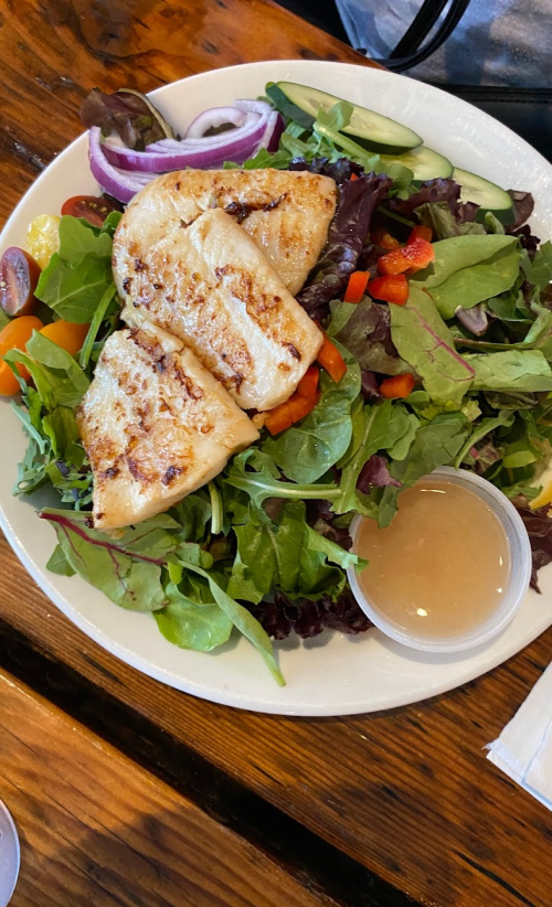 A plate of grilled fish on a bed of mixed greens, topped with tomatoes, cucumbers, and a side of dressing.