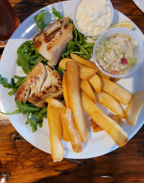 A plate with grilled fish, French fries, a side salad, and dipping sauce on a wooden table.
