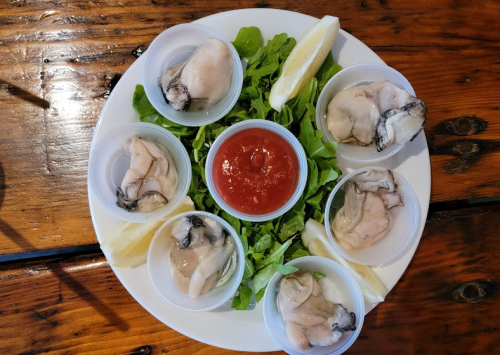 A plate with six cups of oysters on a bed of greens, accompanied by a bowl of cocktail sauce and lemon wedges.