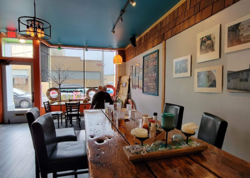 A cozy café interior with a long wooden table, empty chairs, and a view of the street through large windows.