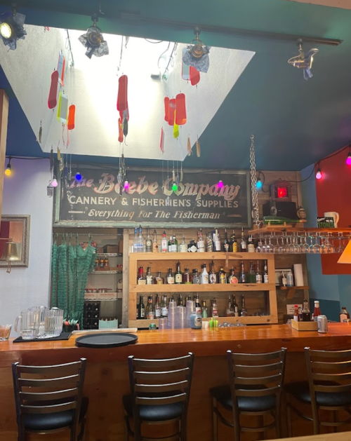 A cozy bar area with a wooden counter, shelves of bottles, and colorful decorations hanging from the ceiling.
