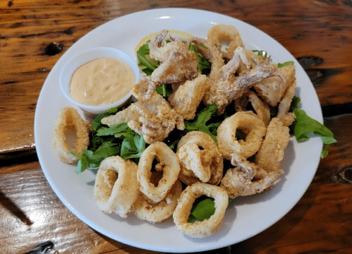 A plate of fried calamari and crispy rings served with a side of dipping sauce and garnished with greens.