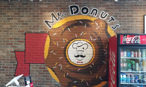 Mural of a donut with a smiling chef's face, labeled "Mr. Donuts," on a brick wall next to a soda vending machine.