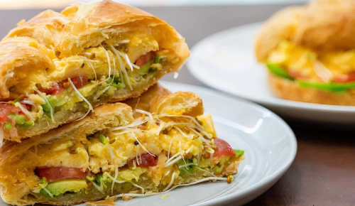Two flaky pastries filled with scrambled eggs, tomatoes, and greens, served on a white plate. A second pastry is in the background.