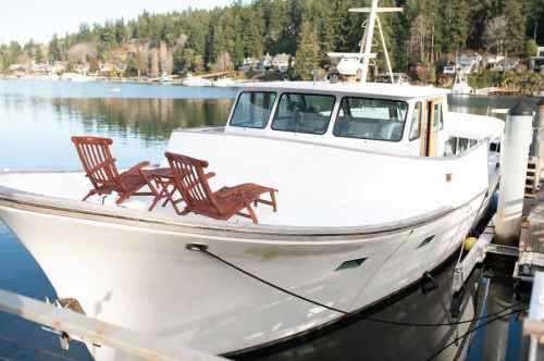 A white boat docked by the water, featuring two wooden chairs on the deck, surrounded by trees and houses in the background.