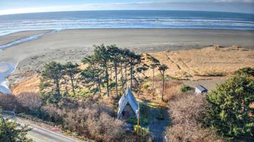 Aerial view of a coastal landscape featuring a unique house surrounded by trees, with a beach and ocean in the background.