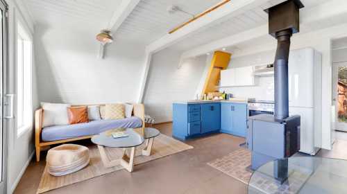 Cozy living space with a blue kitchen, a sofa, coffee table, and a wood stove, featuring bright, natural light.