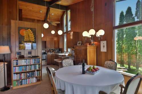 Cozy dining area with a round table, bookshelves, and a modern kitchen, featuring large windows and warm wooden accents.