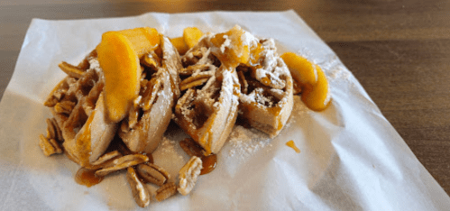 Two waffles topped with pecans, powdered sugar, and slices of peach, served on a white paper.