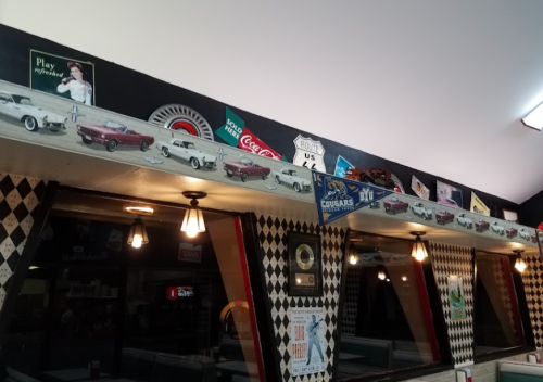 A retro diner interior featuring vintage car decor, Coca-Cola signs, and a checkered wall design.