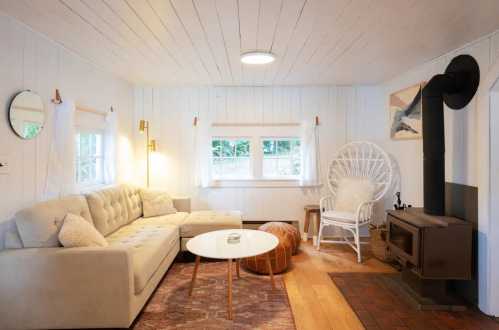 Cozy living room with a light sofa, white walls, a round table, and a wood stove, featuring natural light and warm decor.