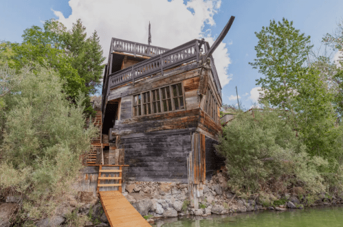 A unique, rustic wooden house perched on a hillside, surrounded by greenery and overlooking a body of water.