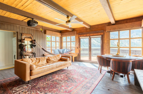 Cozy interior with a leather sofa, wooden beams, large windows, and a dining area overlooking a scenic view.