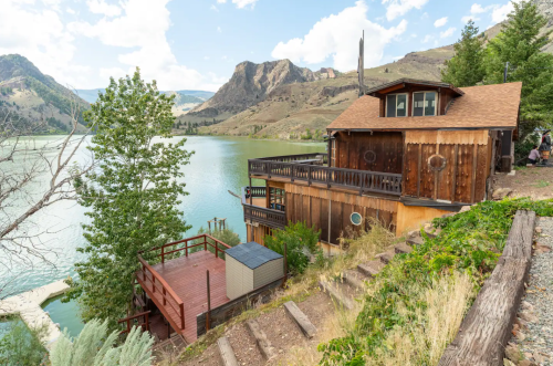 A rustic wooden house by a serene lake, surrounded by trees and mountains under a partly cloudy sky.