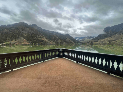 A scenic view from a deck overlooking a calm lake surrounded by mountains under a cloudy sky.