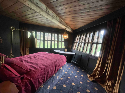 Cozy bedroom with a dark wood ceiling, large windows, a plush bed with burgundy bedding, and a freestanding bathtub.