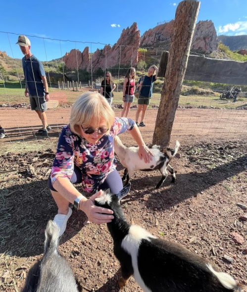 A woman pets a goat while others watch in a scenic outdoor setting with rocky hills in the background.