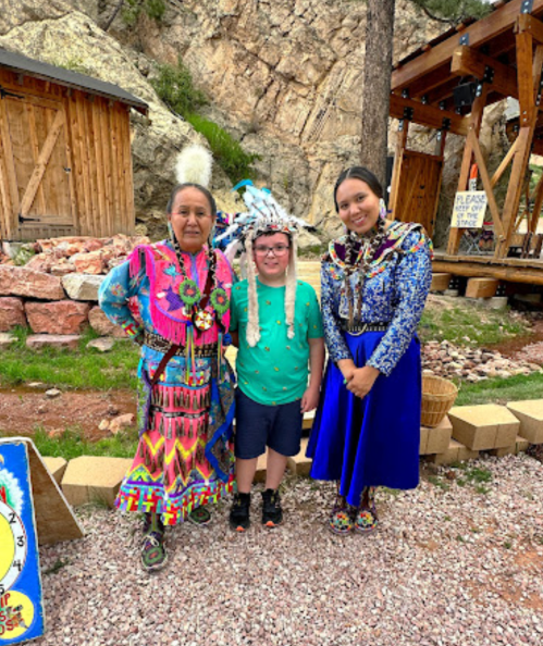 Three individuals pose together outdoors: two women in traditional attire and a child in a colorful shirt, smiling.