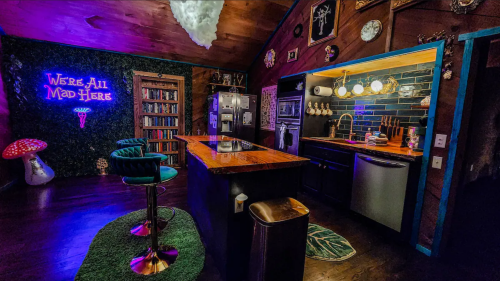 A cozy kitchen with a bar, neon sign, bookshelves, and eclectic decor in warm lighting.