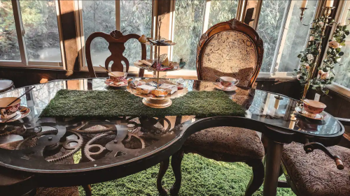 A beautifully set dining table with a glass top, green decor, and elegant chairs, surrounded by natural light.