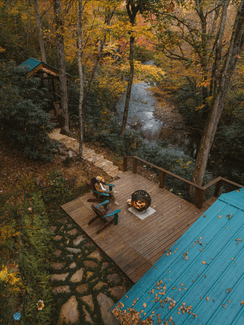 A cozy cabin deck with a fire pit, surrounded by autumn foliage and a winding stream below.