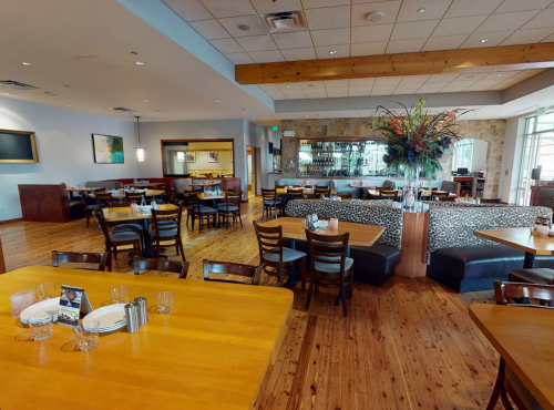 A spacious restaurant interior with wooden tables, chairs, and a bar area, featuring modern decor and a floral arrangement.