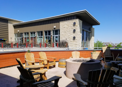 A sunny outdoor patio with wooden chairs and a fire pit, adjacent to a modern building with large windows.