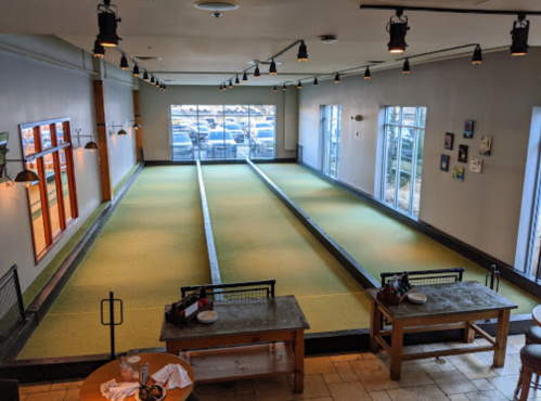 A spacious indoor bocce ball court with green lanes, large windows, and wooden tables in a well-lit setting.