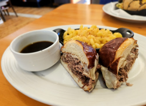 A plate with a pretzel sandwich filled with pulled meat, served with a side of macaroni and cheese and dipping sauce.