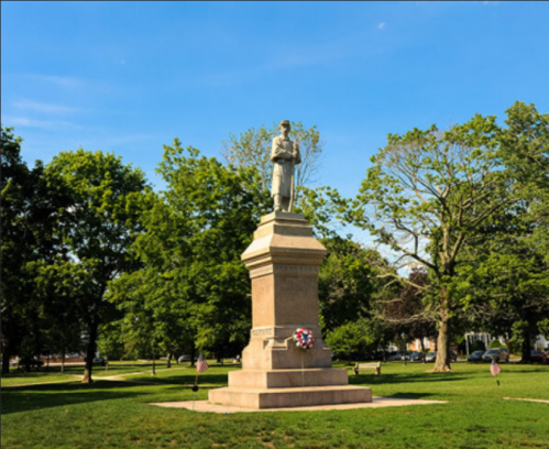 A statue of a soldier on a pedestal surrounded by trees in a park under a clear blue sky.