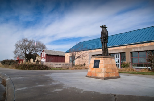 A statue of a cowboy stands in front of the Fort Wallace Museum, with a blue roof and red barn in the background.