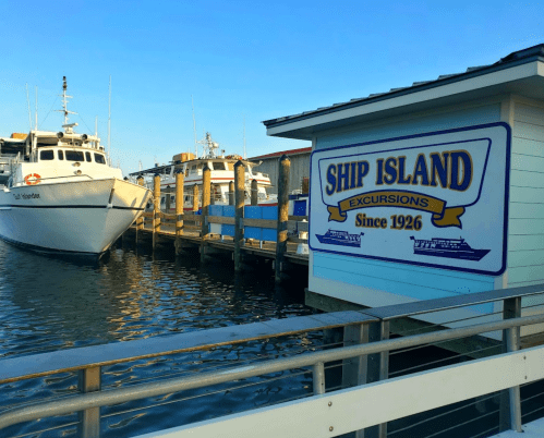 A boat docked at a pier with a sign for Ship Island Excursions, established in 1926, beside calm waters.