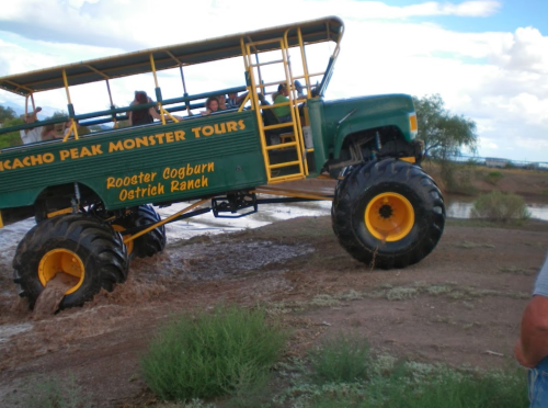 A large, green monster truck with oversized tires navigating through a muddy area, with passengers visible inside.