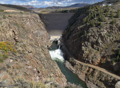 A dam in a rocky canyon, with water flowing from its base into a river below, surrounded by hills and sparse vegetation.